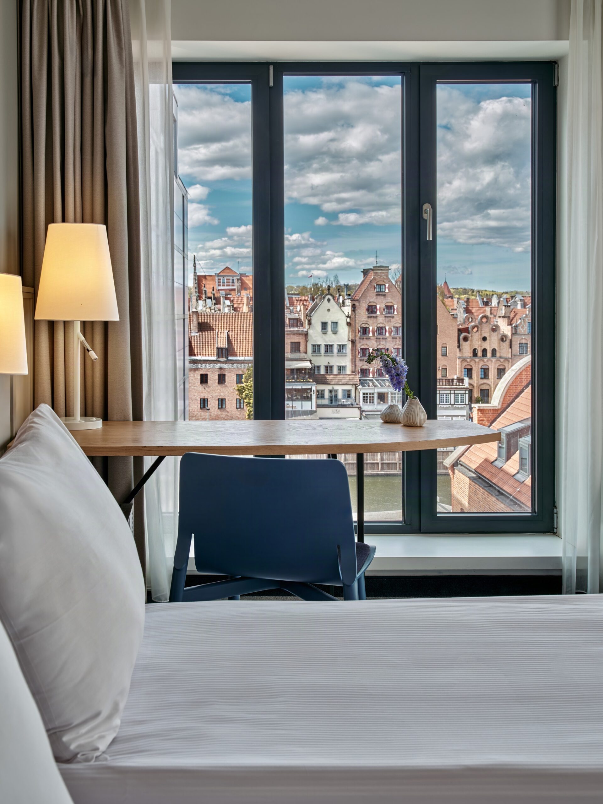 Desk and chair by a large window overlooking red-tiled rooftops and blue sky with clouds in a hotel room.