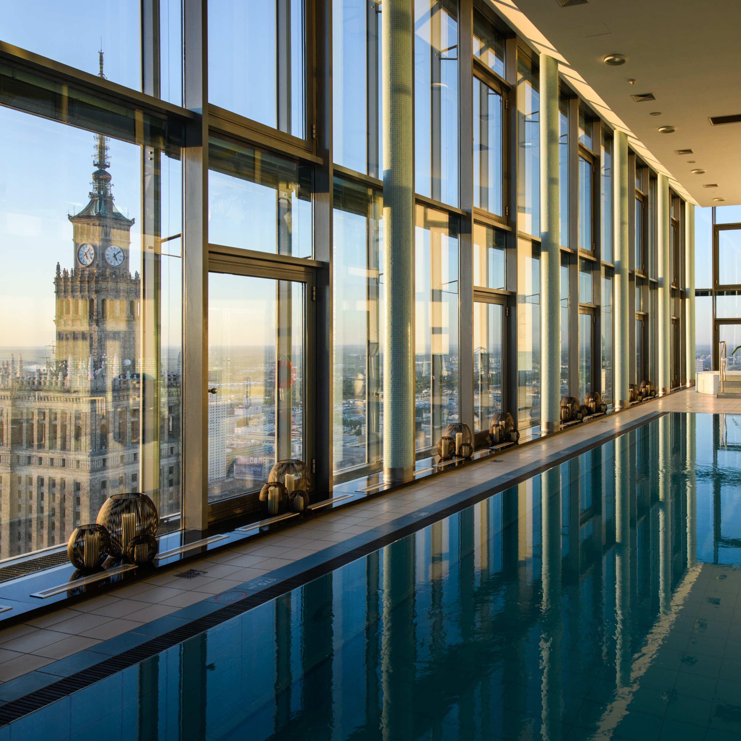 Interior of a modern glass-walled corridor with a reflecting pool and row of lamps, city skyline visible outside at sunset.