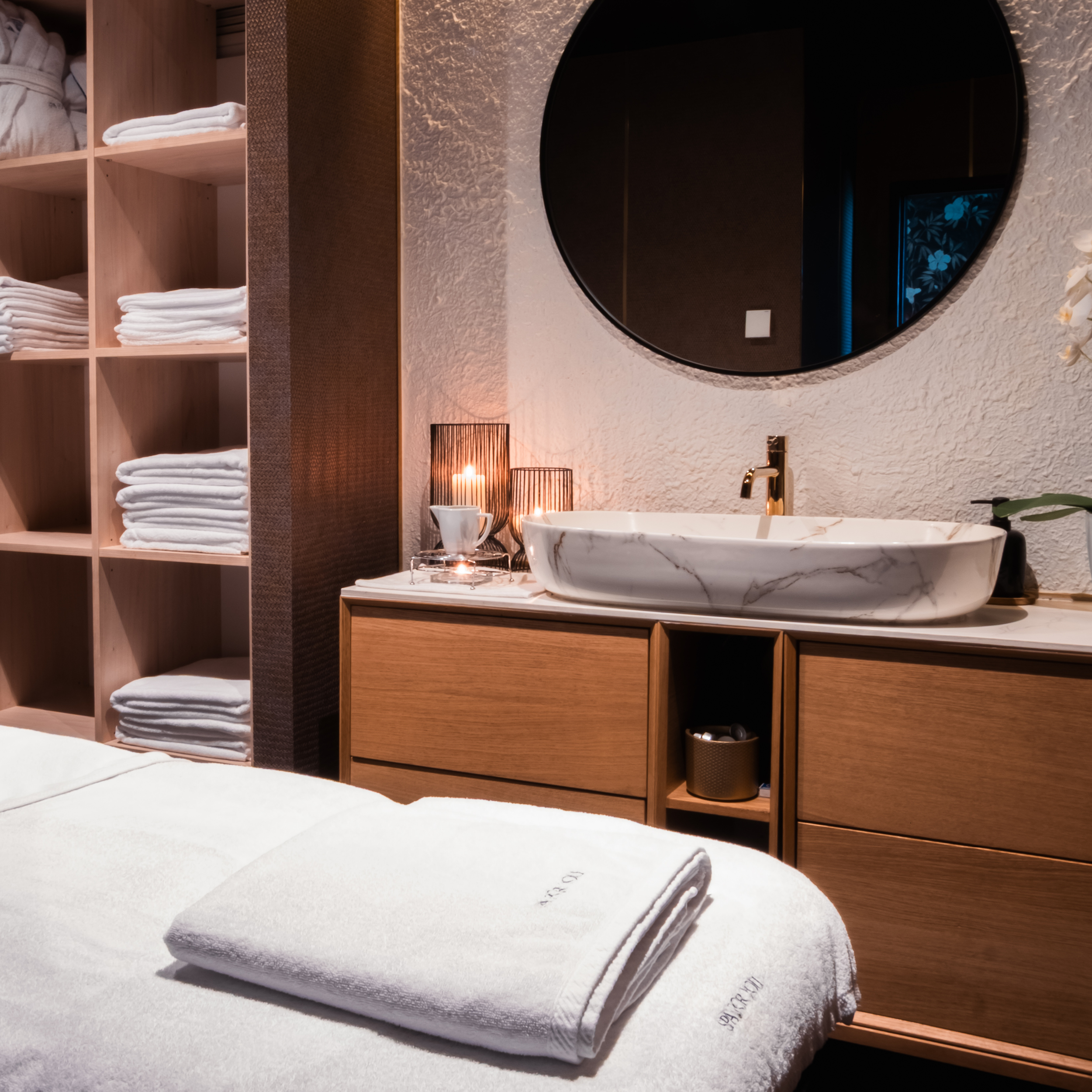 Spa-like bathroom with open wooden shelves holding white towels, a round wall mirror, and a marble sink.