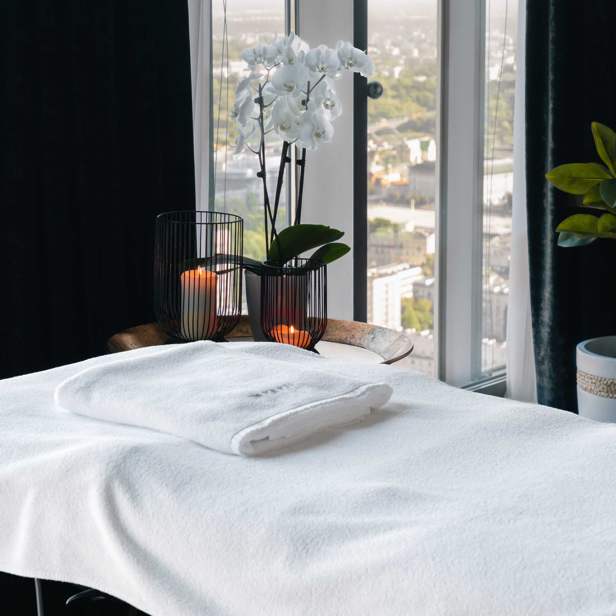 Spa table with folded white towels, lit candles in black wire holders, and a white orchid on the windowsill overlooking a city. (informative)