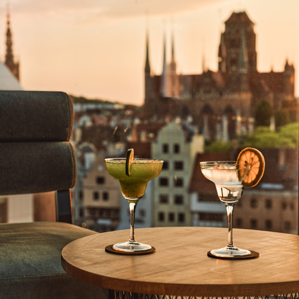 Three cocktails on a round wooden table, sunset city skyline softly blurred in the background.