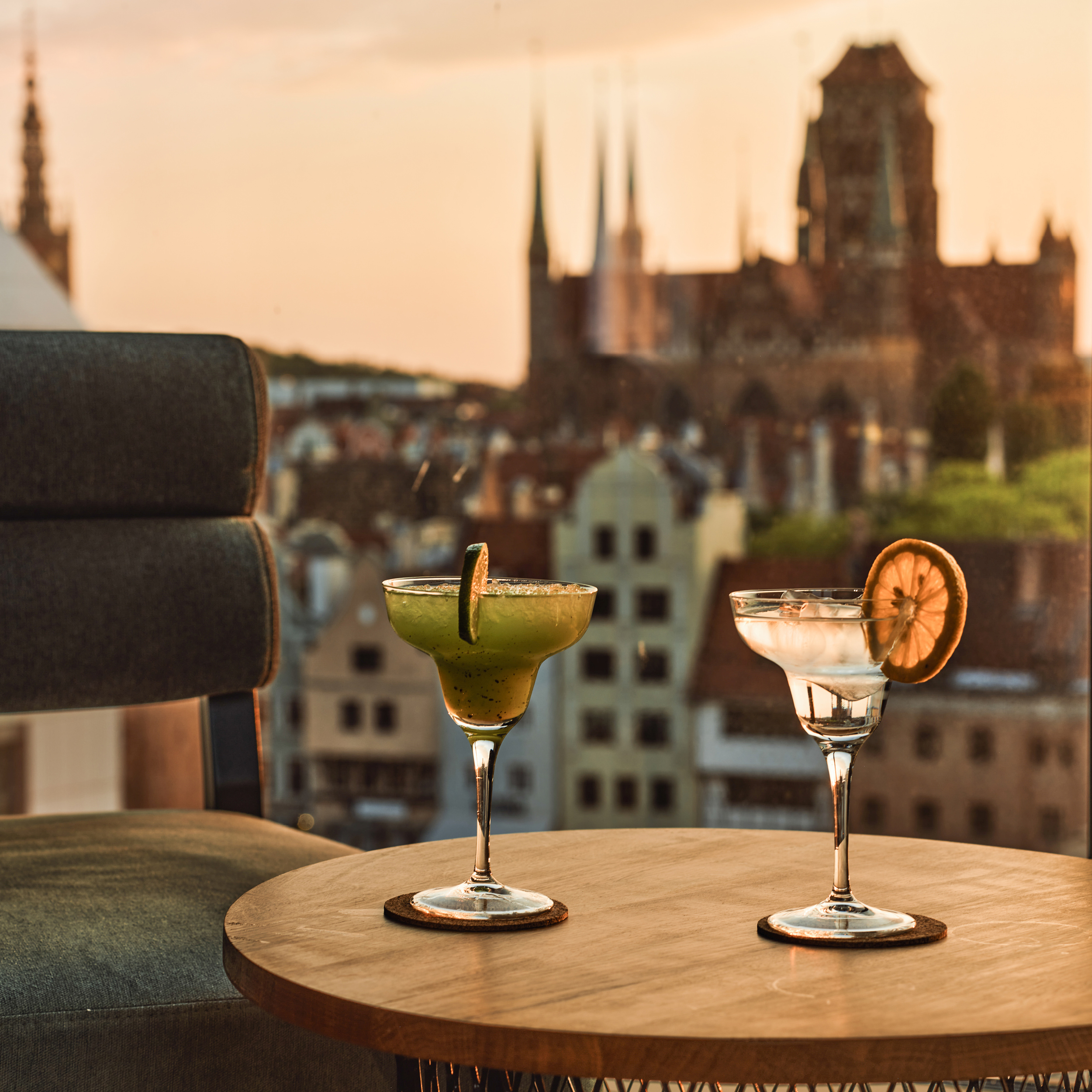 Three cocktails on a round wooden table, sunset city skyline softly blurred in the background.