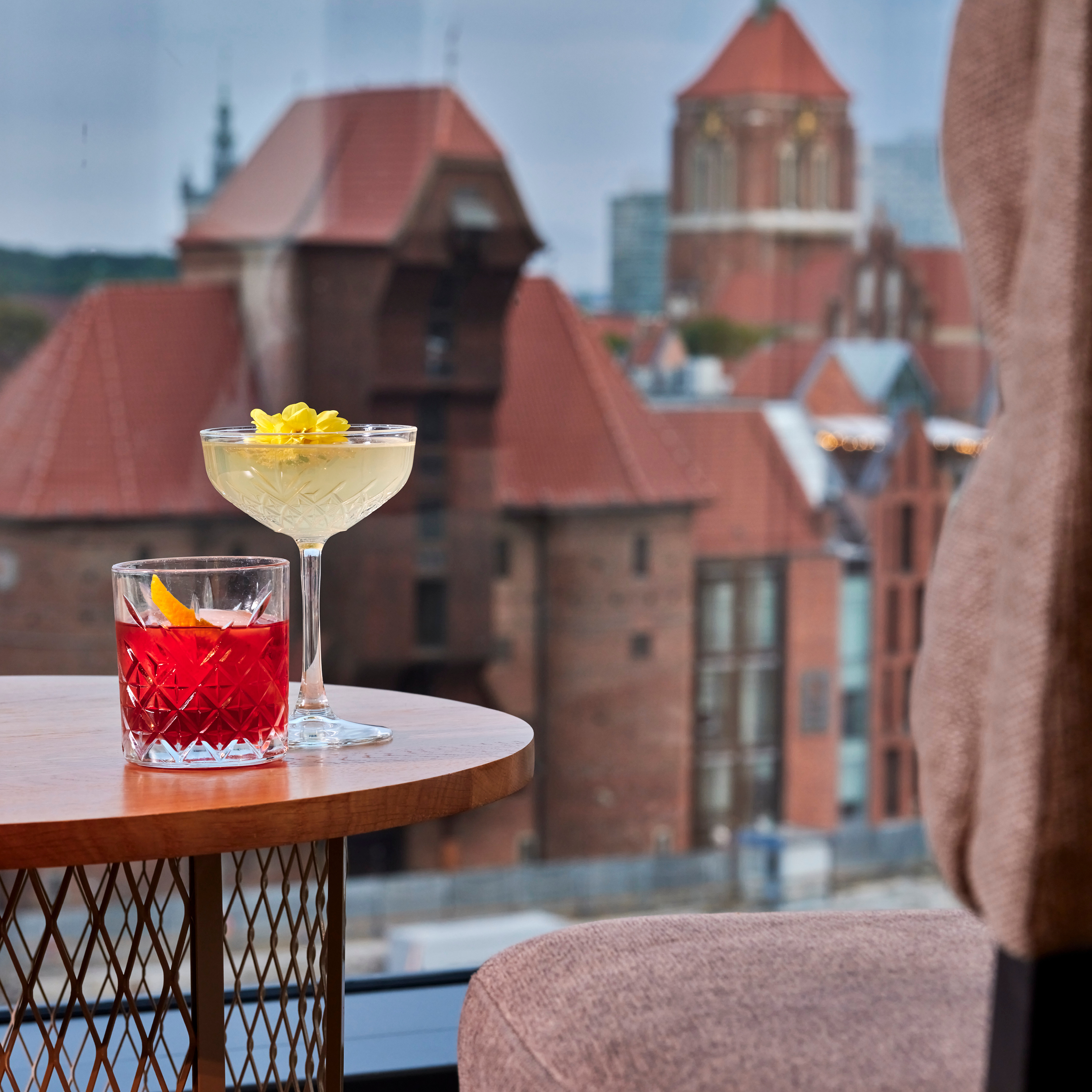 Two cocktails on a rooftop table; a red drink with an orange twist in a faceted glass in the foreground, blurred city rooftops in the background.