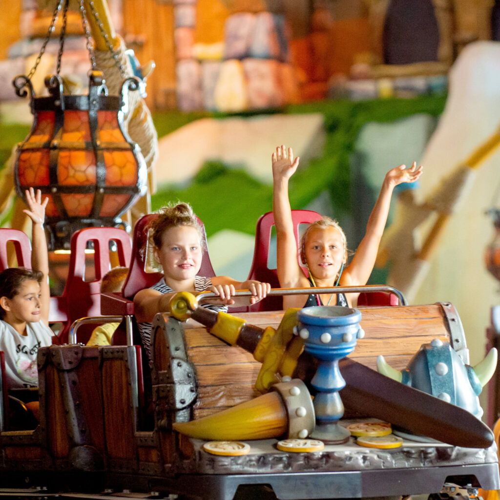 Children on a colorful amusement ride raising their hands in excitement in red seats under warm lights.