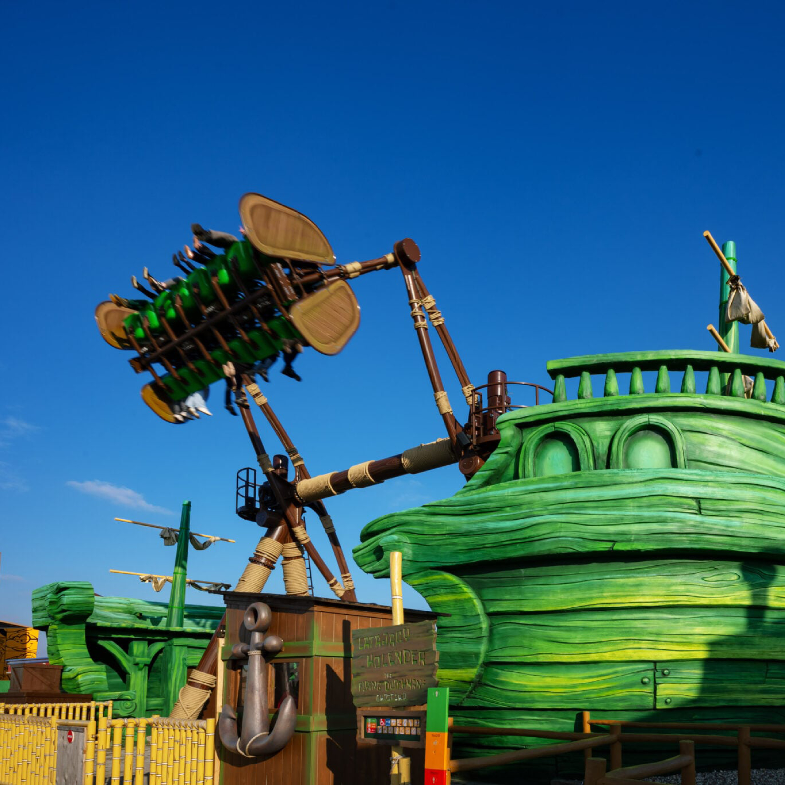 Dragon-themed amusement ride with green planks and brown supports against a clear blue sky; riders suspended on a track above green boat hulls.