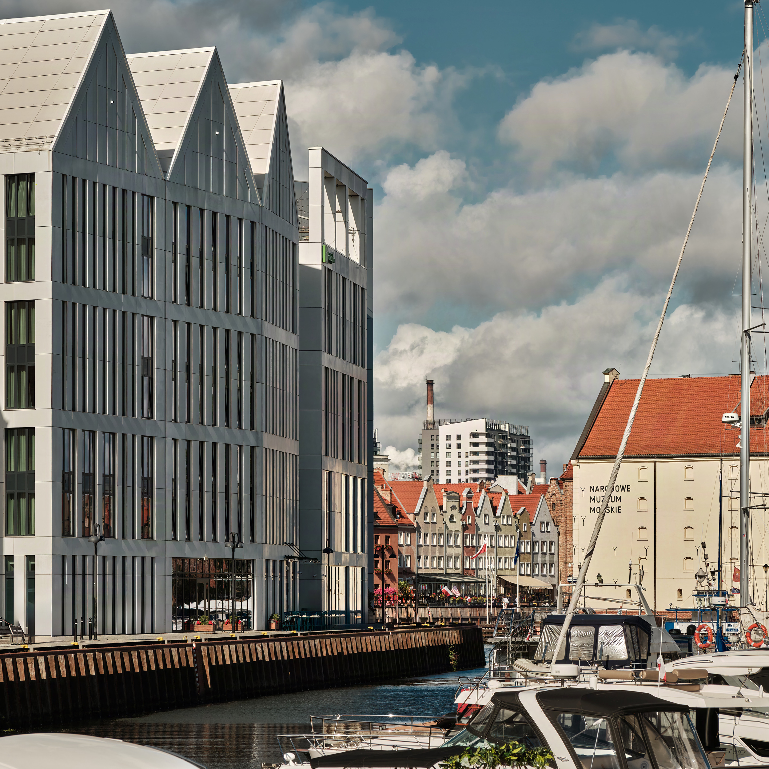 Modern white glass-fronted building along a marina with boats docked in the foreground and red-roofed town buildings in the distance under a cloudy sky