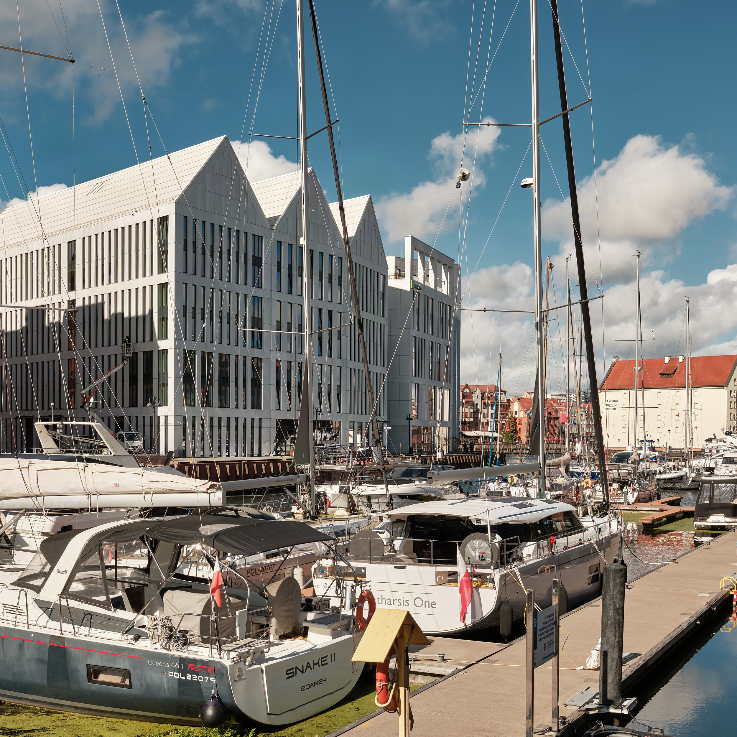 Marina with moored yachts and tall masts beside a modern white building under a blue sky at the harbor.