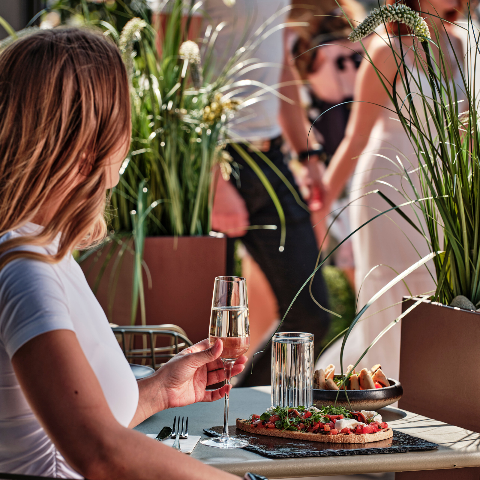 Woman in a white shirt holds a flute of pink drink at an outdoor table, while plates of bruschetta and bread sit nearby among tall plants.