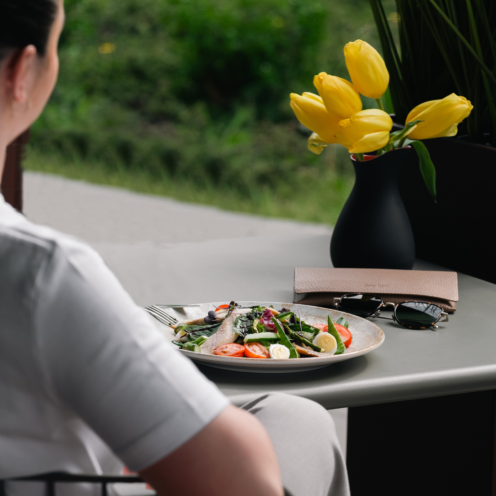 Person dining outdoors at a table with a colorful salad, yellow tulips in a black vase, and sunglasses on the table in view.