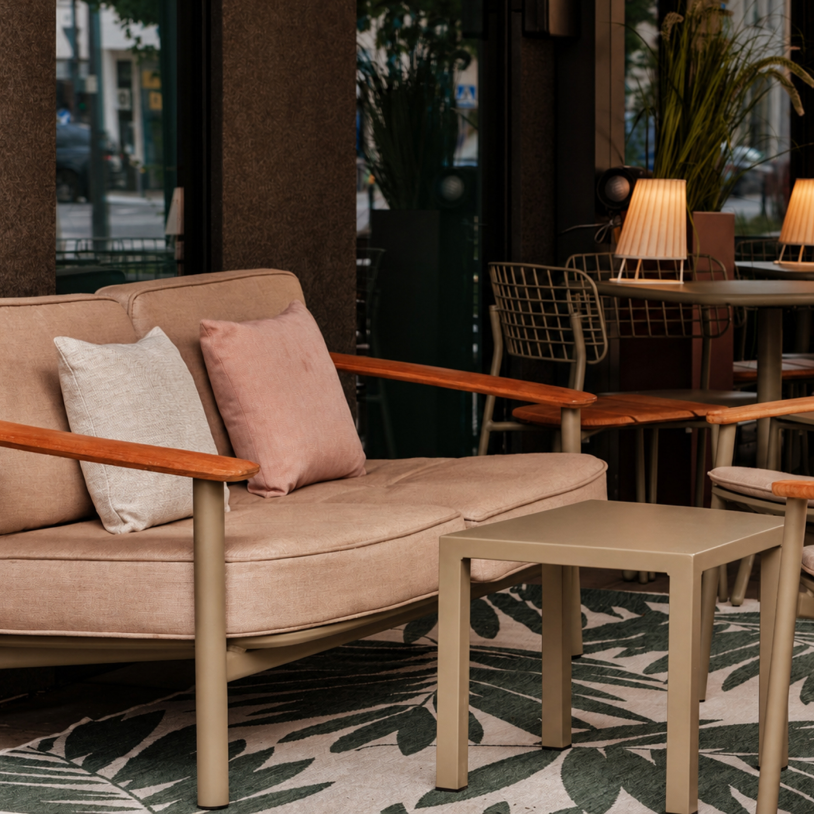 Outdoor cafe seating with a beige cushioned sofa, pale pillows, and wooden armrests; warm lamps on nearby tables.