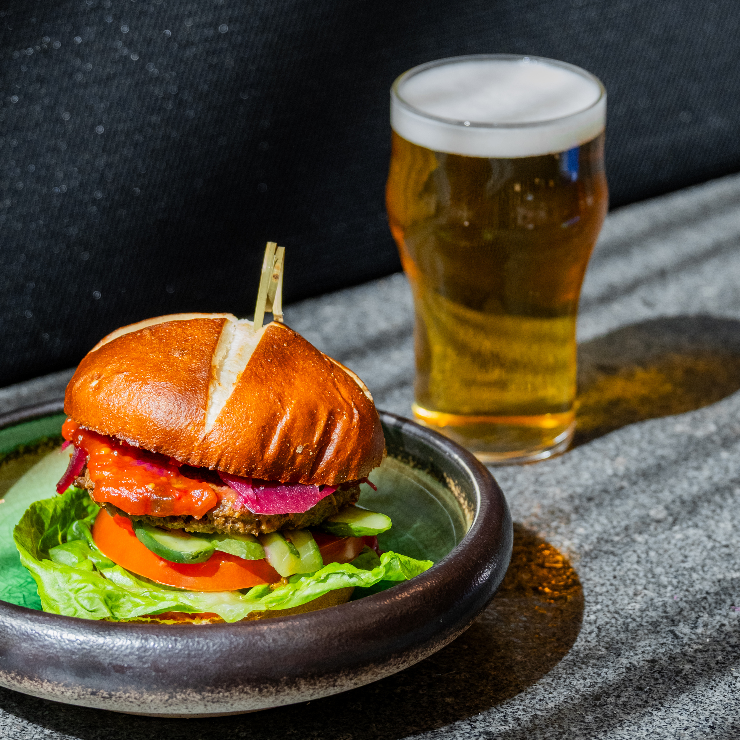 Close-up of a stacked burger with lettuce, tomato, red onions and a glossy bun on a plate; a glass of beer appears in the background.