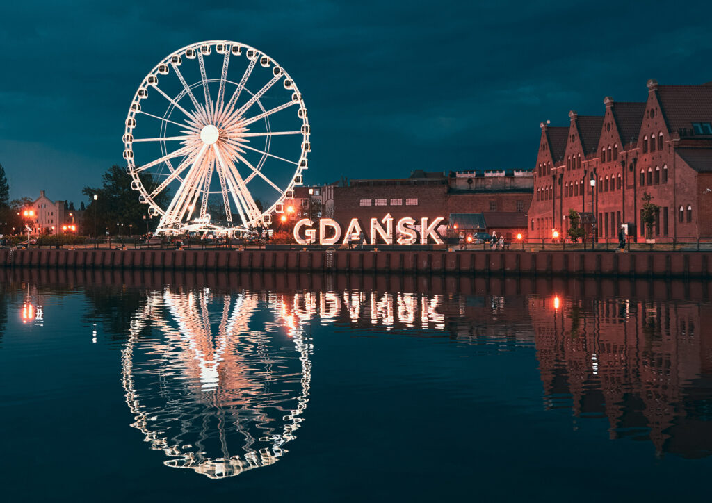 Lit Ferris wheel at the Gdańsk waterfront with brick warehouses and a glowing 'GDAŃSK' sign, reflected in calm water.
