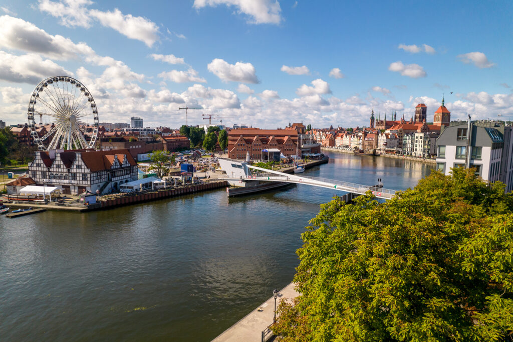 Riverside town with a large Ferris wheel, red-brick buildings, and a pedestrian bridge over a calm river under a blue sky.