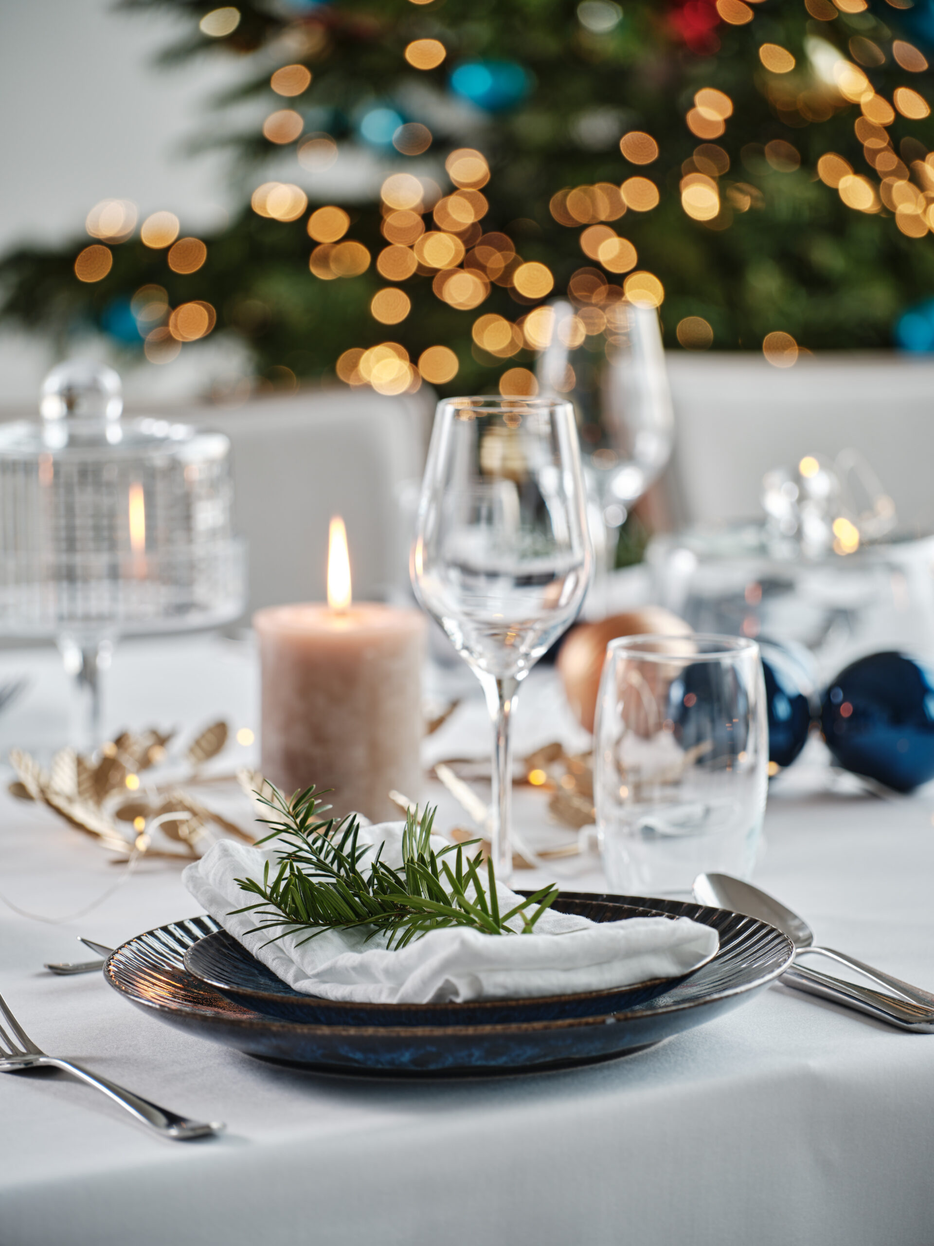 Elegant holiday table setting with a dark plate, white napkin and sprig, candle, and blurred Christmas lights in the background.