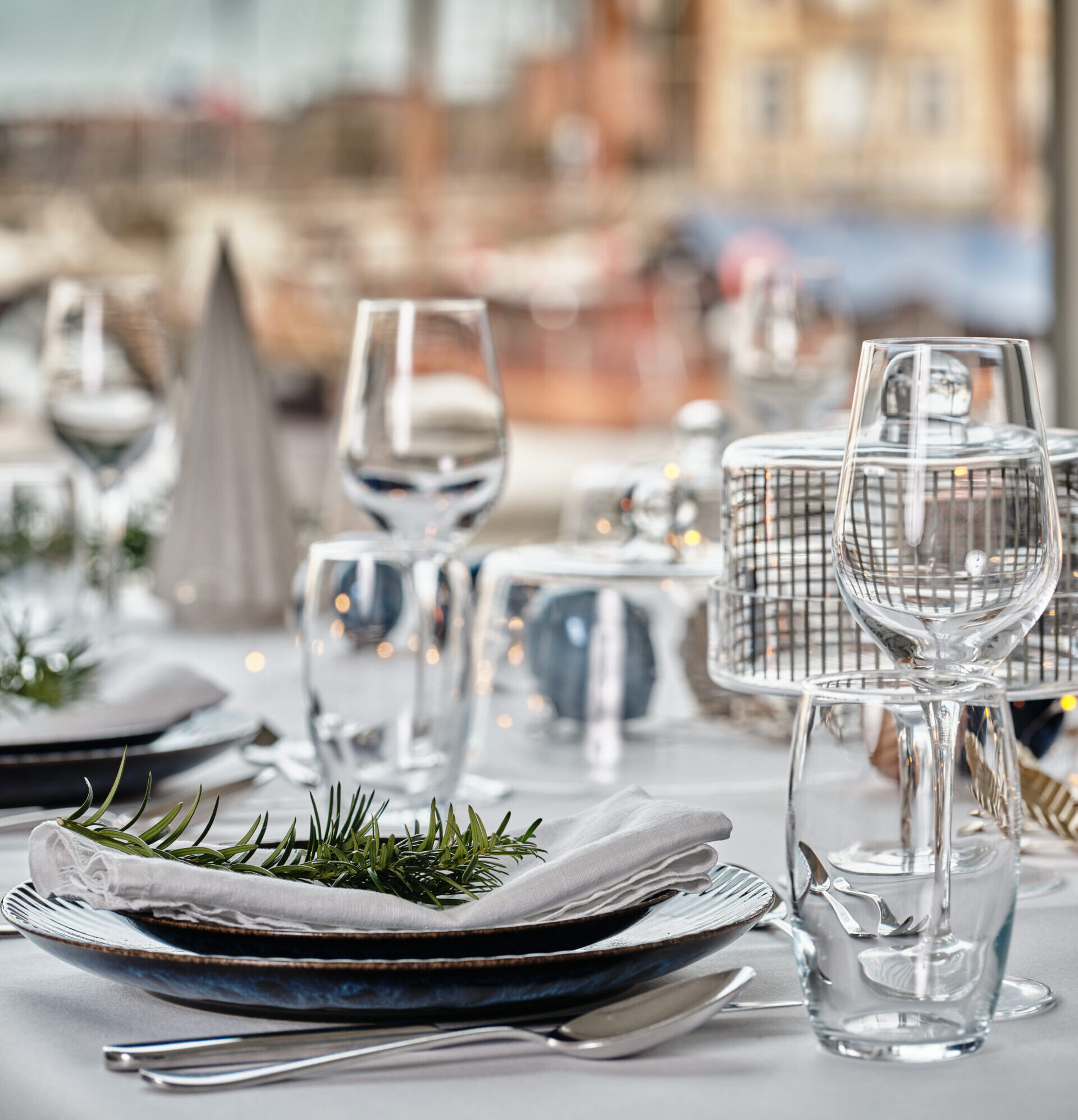 Table setting for dinner with a folded napkin and rosemary sprig on a stack of plates, plus wine glasses in the foreground.