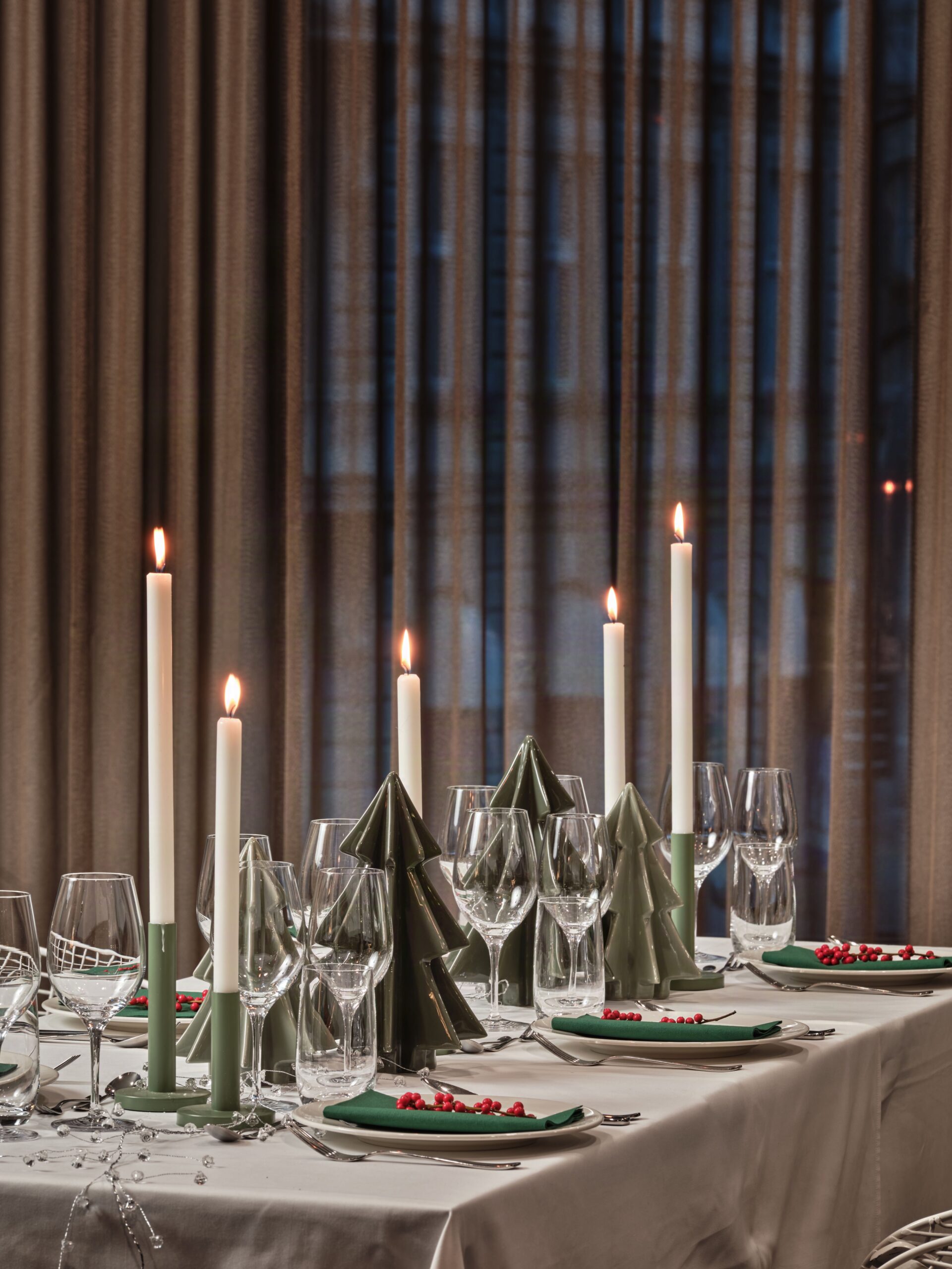 Elegant dining table set for a formal event with tall white candles, green folded napkins, crystal wine glasses, and red berry accents on a white tablecloth.