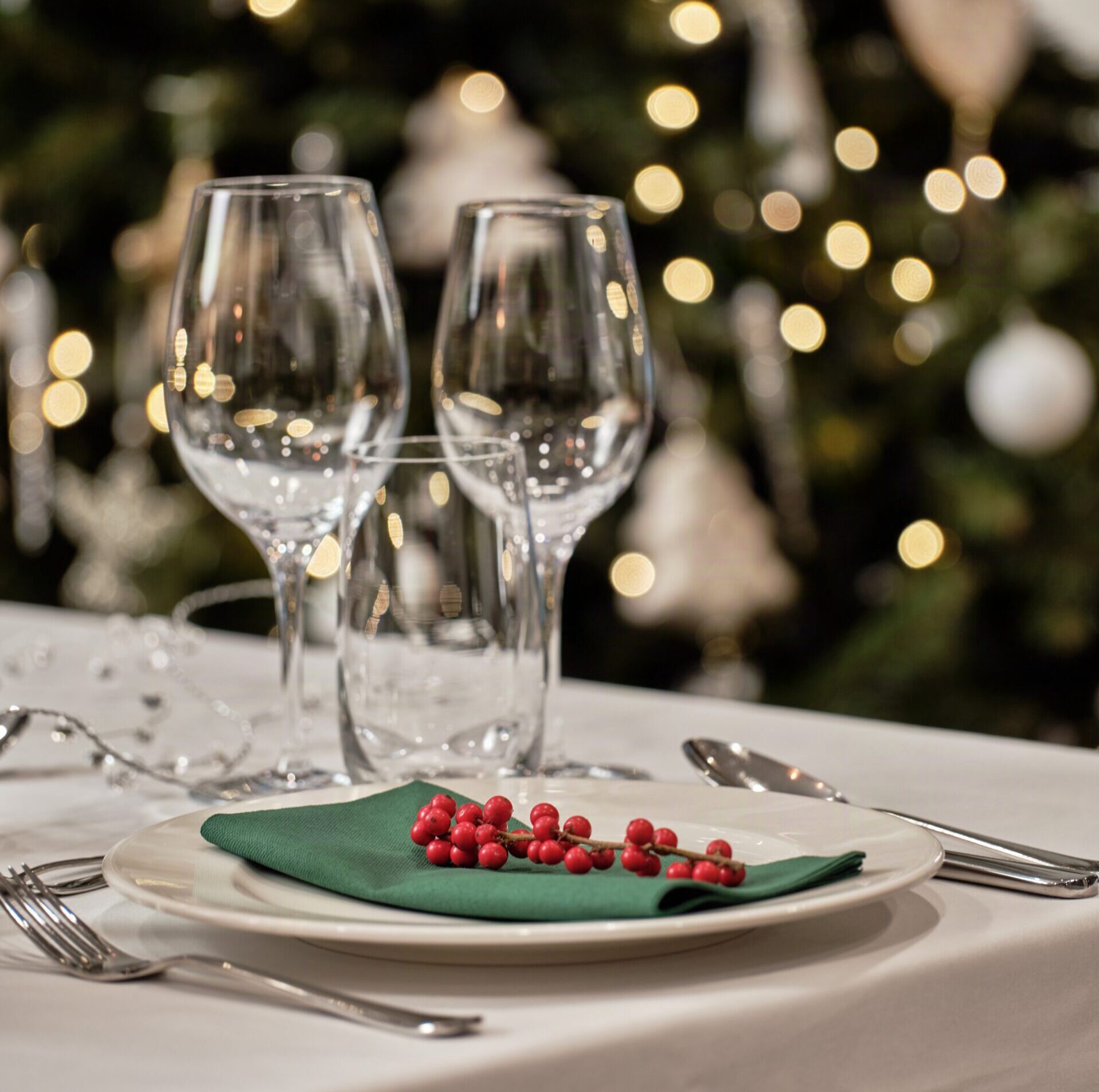 Elegant table setting with two wine glasses, a white plate, a green napkin, and red berries; warm Christmas lights in the background.