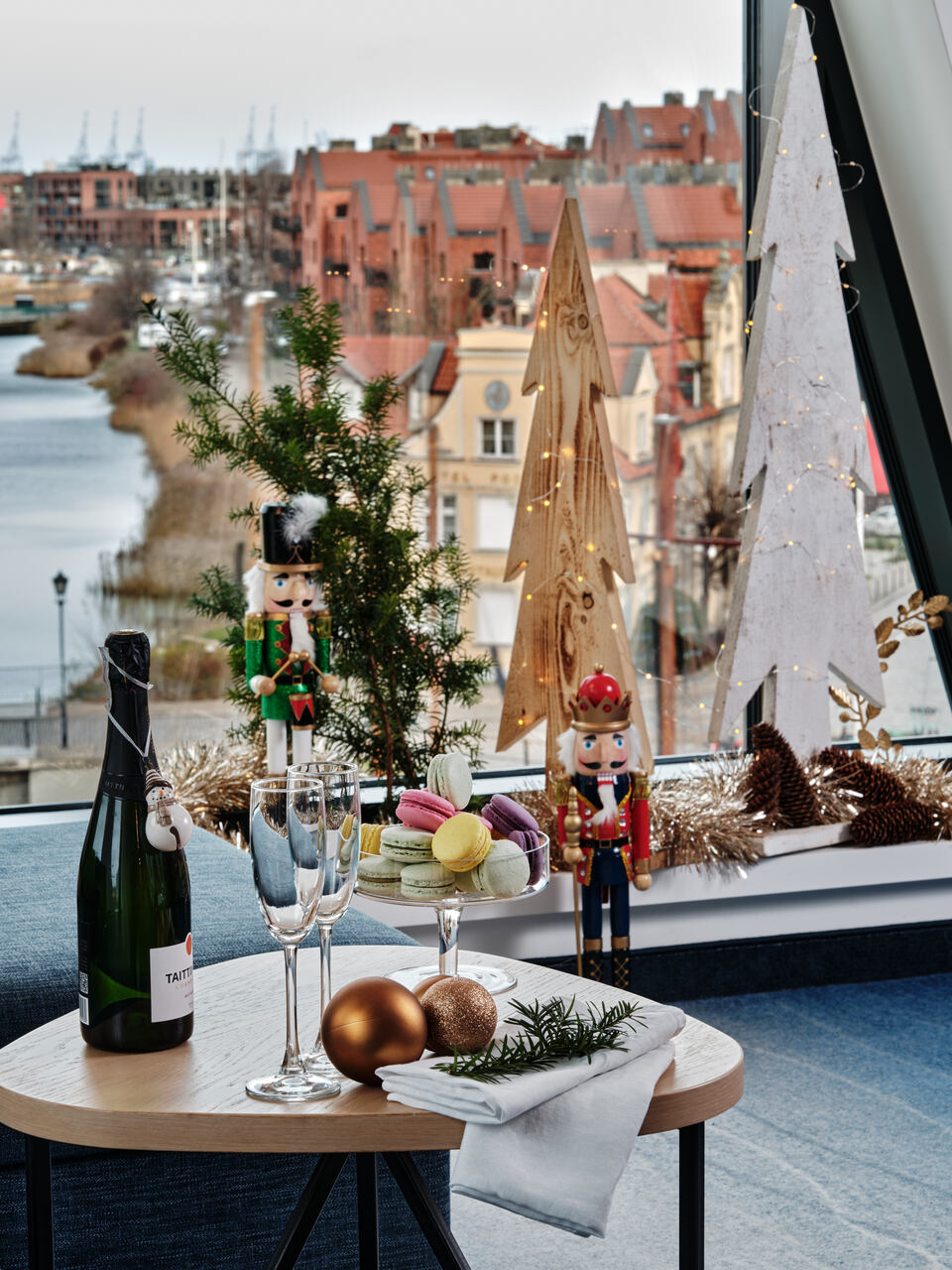 Table by a window with champagne, macarons, nutcrackers, and twinkling fairy lights overlooking a riverside village, festive setup.