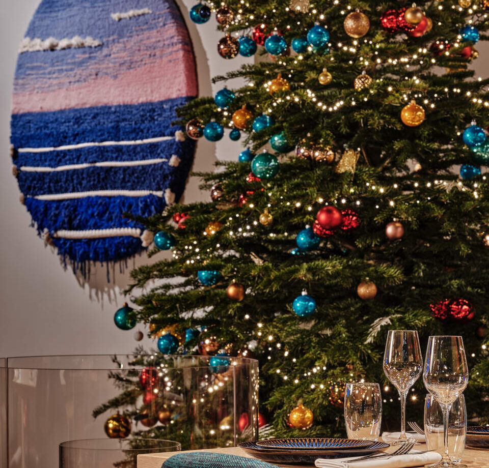 Christmas tree with colorful ornaments and warm string lights beside a dinner table set with plates, glassware, and a lit fireplace in the background, creating a festive scene.