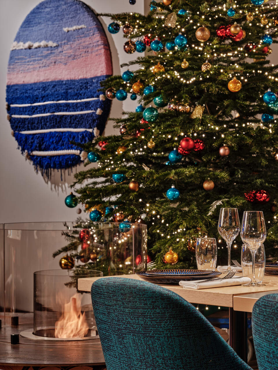 A dining table set for a holiday meal beside a lit Christmas tree with colorful ornaments and string lights, plus a teal chair and a cozy fireplace nearby.
