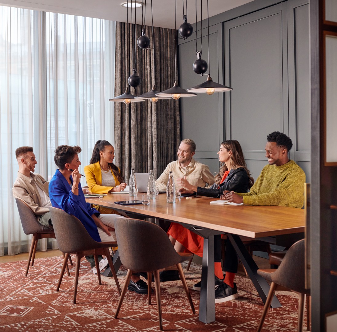 Six professionals of diverse backgrounds sit around a wooden conference table in a modern room, smiling and talking during a meeting.